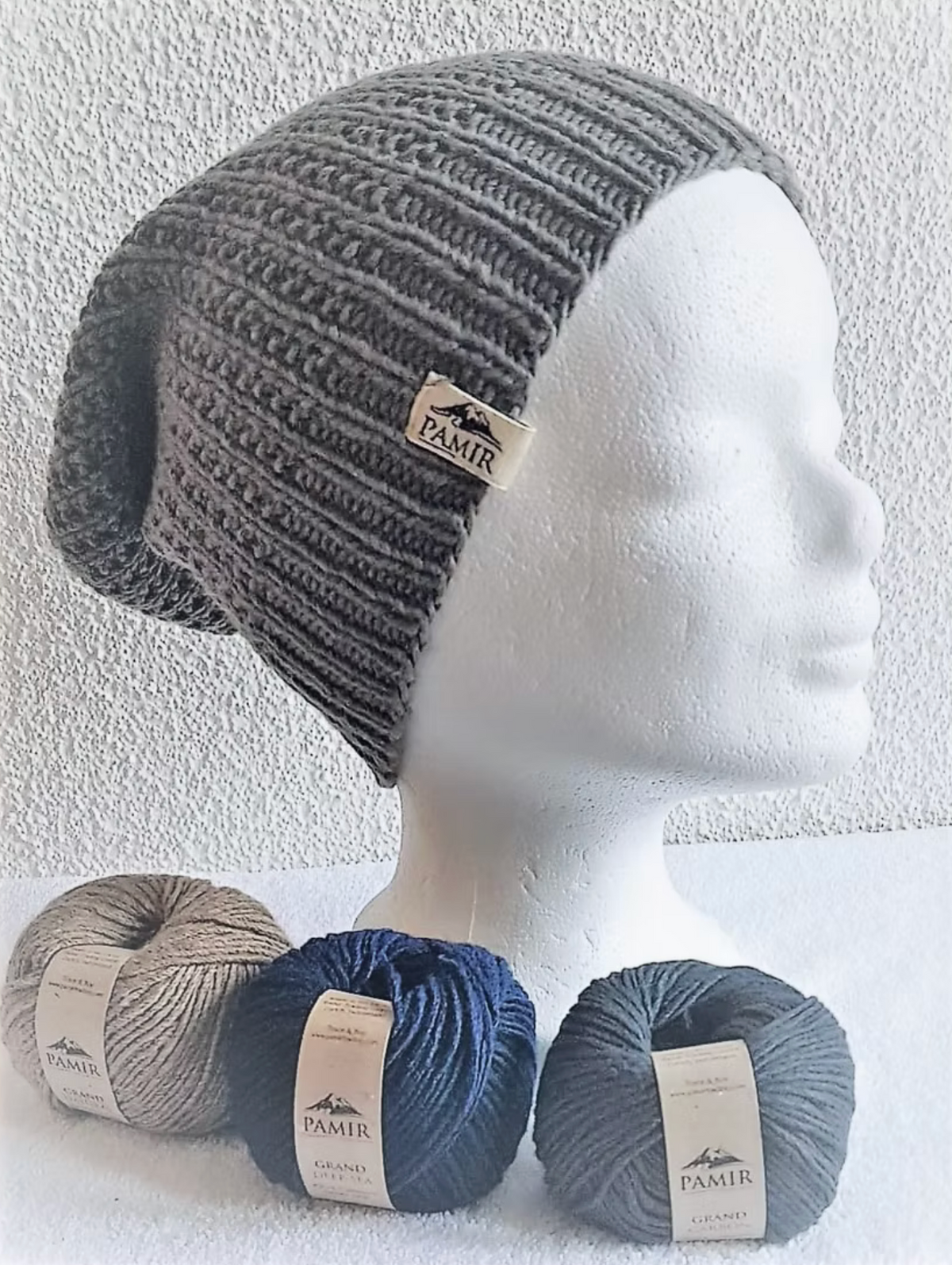 Graue Beanie N#4 auf einem Kopfmodell neben zwei Knäueln GRAND Kaschmir